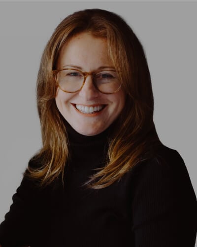Trisha Donkers, with long light brown hair and glasses, smiles at the camera. She is wearing a black turtleneck sweater and is set against a plain gray background.
