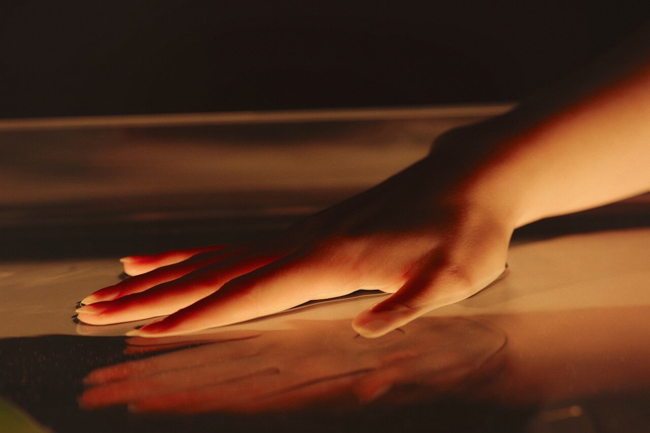 A hand softly pressing on a smooth, reflective surface with warm lighting highlighting the fingers and casting a gentle shadow.